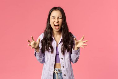 Portrait of aggressive young bothered girl losing temper, screaming furious and angry grimacing, yelling at person with hate and aggression, squeeze hands into fists, pissed-off, pink background
