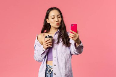Güzel görünümlü bir kadın blogcunun portresi, yaşam tarzı internet etkileyicisi en sevdiği kafeden alınmış kahveyle selfie çekiyor, cep telefonu tutuyor, öpücük ve kapalı gözler için somurtuyor.