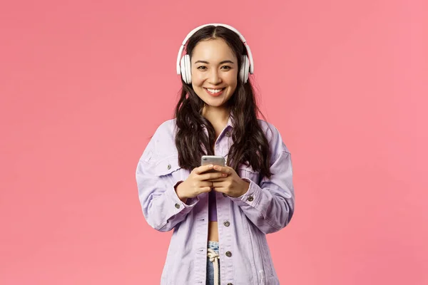 Portrait of attractive modern and stylish brunette korean, asian girl in headphones using mobile phone, listening music, made new playlist for going out, standing pink background