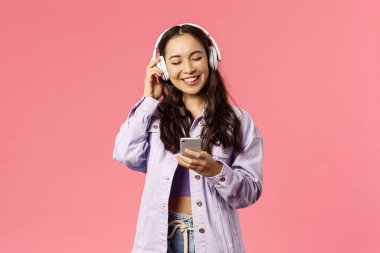 Tarz sahibi, çekici ve mutlu, gülümseyen Asyalı bir kızın portresi kulaklıkla favori şarkıları dinlerken, gözleri kapalıyken, gülerken, müzik kanalı ya da komik bir podcast bulurken, cep telefonunu tutarken.