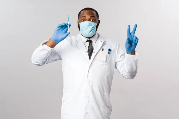 Covid-19, traitement du coronavirus et concept de laboratoire. Heureux médecin afro-américain traitant les gens à l'hôpital, montrer un signe de paix et tenir la seringue avec un vaccin bleu, porter un masque facial — Photo