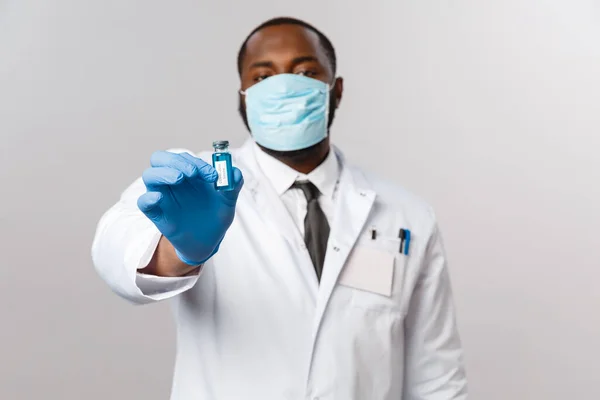 Covid19, πανδημία και υγειονομική περίθαλψη έννοια. Σοβαρός αφροαμερικανός γιατρός με λευκό παλτό, γάντια λάτεξ και ιατρική μάσκα προσώπου που δείχνει νέο εμβόλιο από το ξέσπασμα του κορωναϊού, θεραπεύει ανθρώπους — Φωτογραφία Αρχείου