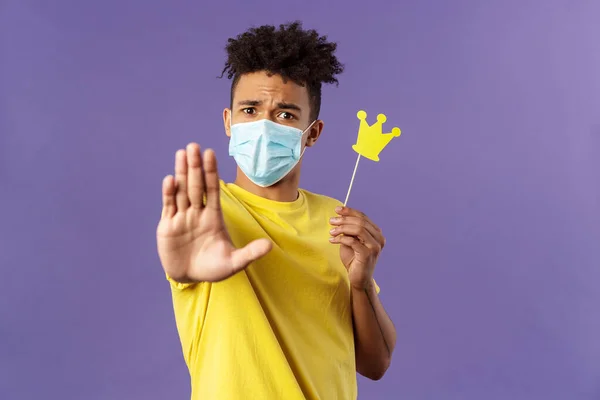 Close-up portrait of young hipster guy in medical mask stretch hand forward stop gesture, holding small paper crown, demand stay away, keep 1.5 meter distance, avoid social contact during quarantine