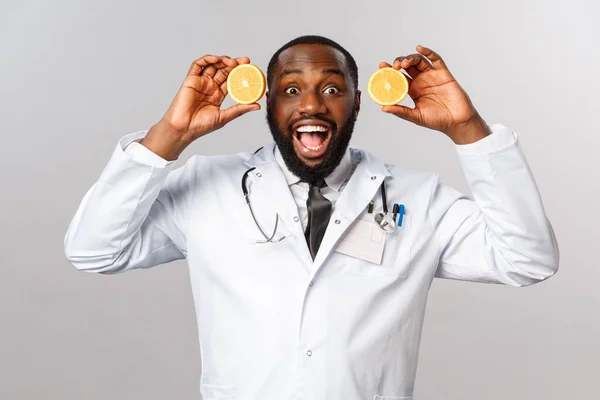 Здравоохранение, здоровое питание и концепция заболеваний. Возбужденный красивый африканско-американский врач в белом халате, показывая кусочки апельсинов и улыбаясь весело, совет съесть больше пищи состоят из витамина С, фрукты — стоковое фото