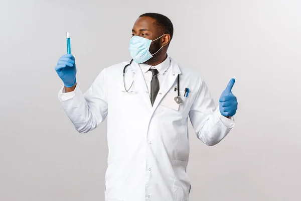 Covid-19, behandeling van coronavirus patiënten en laboratoriumconcept. Tevreden Afrikaans-Amerikaanse dokter vond virusvaccin uit, verheugd kijkend naar flacon, reageerbuis met blauwe vloeistof, duim omhoog — Stockfoto