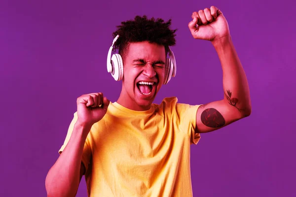 Primer plano retrato de alegre, joven bailarín feliz levantar la mano cantando, cerrar los ojos y sonriendo optimista como escuchar canción impresionante en los auriculares, disfrutando de la música, fondo púrpura —  Fotos de Stock