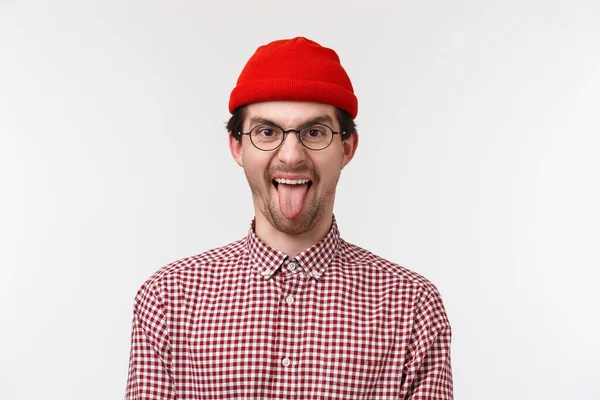 Close-up portrait funny cute bearded hipster guy in red beanie and glasses, smiling showing tongue playfully, having fun acting childish standing white background joyful — 스톡 사진