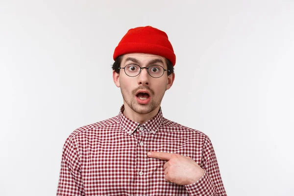 Närbild porträtt förvånad ung hipster kille i glasögon och mössa stirrar med misstro, pekar sig obeslutsam, vet inte om personen på riktigt nämnde honom, stående vit bakgrund — Stockfoto
