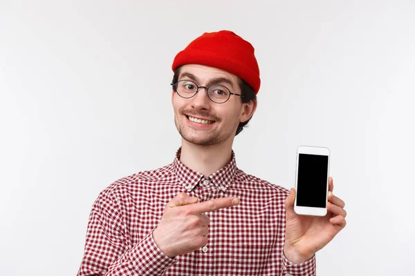 Conceito de tecnologia e pessoas. Retrato close-up jovem homem moderno com barba em gorro e óculos, mostrando sua nova aplicação que ele programou, apontando para a tela do telefone móvel, sorrindo satisfeito — Fotografia de Stock