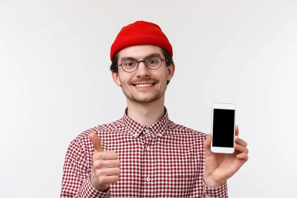 技术和人的概念。特写满足了相貌英俊、留着胡子、戴着眼镜、头戴红色贝尼帽的男人的要求，并在手机广告中展示了大拇指，站在白色背景上 — 图库照片