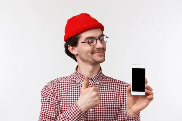 Conceito de tecnologia e pessoas. Close-up retrato satisfeito feliz jovem hipster homem em gorro vermelho e óculos, olhando satisfeito na tela do telefone móvel, fazer thumb-up aprovar gadget smartphone — Fotografia de Stock