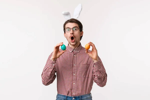 Traditions, fêtes religieuses, concept de célébration. Jeune homme joyeux drôle avec barbe dans les lunettes, porter des oreilles de lapin mignon et tenir deux œufs peints, jouer au jeu le jour de Pâques, fond blanc — Photo