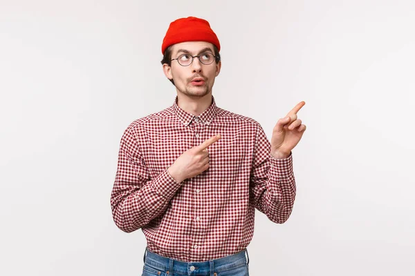 Ενδιαφέρουσα ματιά. Περίεργος νεαρός καυκάσιος με γενειάδα σε κόκκινο σκούφο και γυαλιά, κρυφοκοιτάζει στην πάνω δεξιά γωνία με ενδιαφέρον και περιέργεια, στέκεται λευκό φόντο — Φωτογραφία Αρχείου