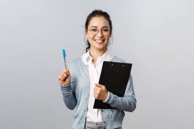 Eğitim, öğretmenler, üniversite ve okul konsepti. Mutlu genç bayan çalışan, ofis çalışanı, not alıyor, elinde kalem tutuyor, mutlu gülümsüyor, gri arka planda duruyor.