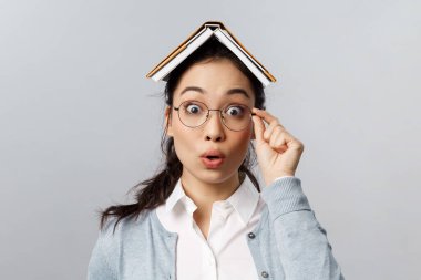 Eğitim, üniversite ve halk konsepti. Şaşırmış Asyalı bir kadının yakından çekilmiş portresi, hayranlıkla baktı ve merak etti, gözlük takıyordu, kafasında kitap tutuyordu, gri arka planda duruyordu.