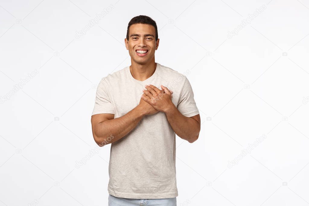 Guy feeling touched and flattered receiving praises. Charming hispanic young man in t-shirt, press hands to heart, smiling and laughing carefree, express gratitude, white background