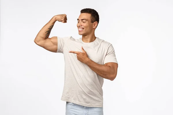 Satisfeito, orgulhoso e assertivo jovem brasileiro com músculos fortes, braço levantado tenso, sorrindo e apontando bíceps orgulhosamente, encantado com seu próprio corpo perfeito, treino no ginásio, mostrando resultados do esporte — Fotografia de Stock