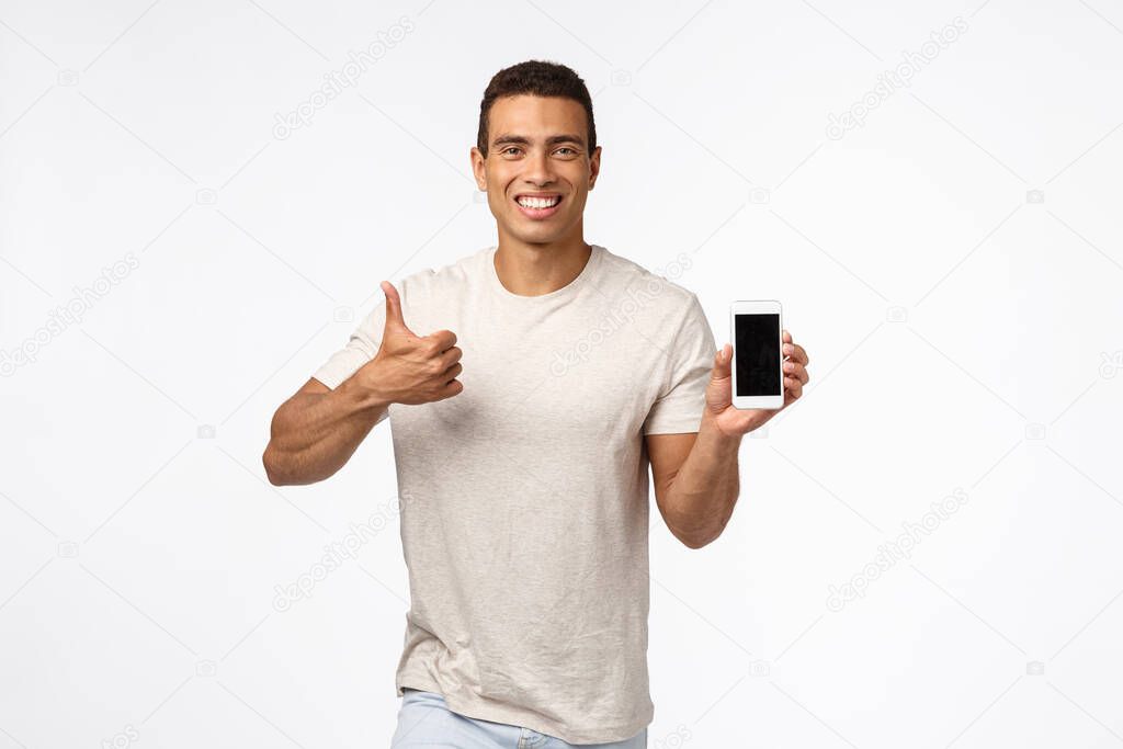 Satisfied, cheerful good-looking sexy young man in t-shirt, showing thumbs-up in approval or like gesture, smiling nod in agreement, recommend app, holding smartphone, introduce gadget