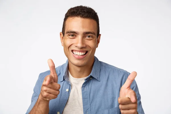 Cheeky, bel homme charismatique hispanique en chemise bleue sur t-shirt, pointant du doigt pistolets caméra, souriant amusé, encourager quelqu'un, rencontrer une personne avec un mouvement de salutation amical, fond blanc — Photo