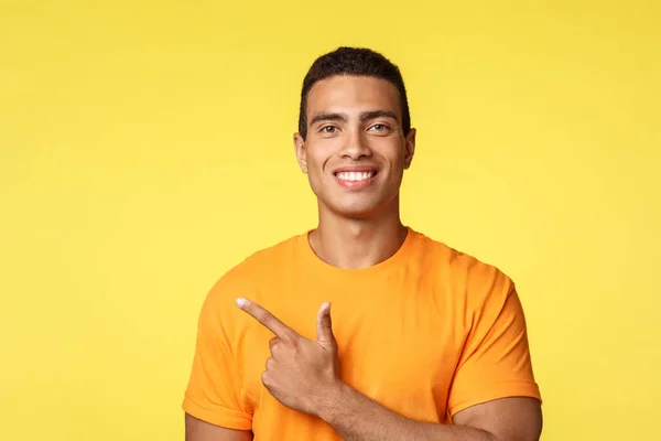 Primer plano joven masculino sonriente, hombre feliz en camiseta, apuntando a la izquierda, gestos y sonriendo orgulloso, mostrando promoción venta o la mejor opción, dar consejos, recomendar tienda, soporte fondo amarillo — Foto de Stock