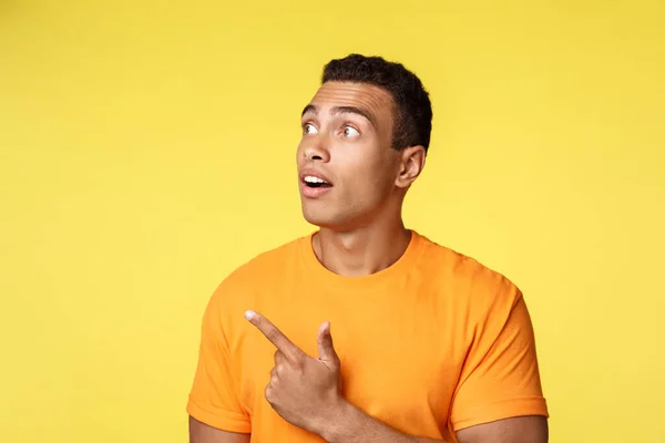 Impresionado, se preguntó joven guapo chico milenario en camiseta, jadeando sorprendido, mirando fascinado y divertido, apuntando a la izquierda en algo interesante, bandera y concepto de promoción, fondo amarillo — Foto de Stock