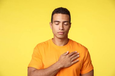 Barışçıl ve umut dolu genç yakışıklı adam, elini kalbine koy, gözlerini kapat ve dua et veya en sevdiklerini düşün, hatırla anı, yemin et, söz ver, sadık ol, sarı arka planda dur