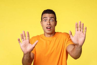 Adam korkmuş, paniklemiş, teslim olmuş, sakin biri olmaya çalışıyor. Ateş etmeyin, nefes nefese kalmayın, endişeli ve endişeli bir şekilde geri çekilin, avuç içinizi kaldırın, sarı arka planda durun, diyor.