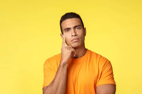 Pensive, serious-looking handsome guy in t-shirt look strict and thoughtful camera, touch temple as trying think, make-up plan, decide, have assumption, standing indecisive yellow background — Stock Photo, Image