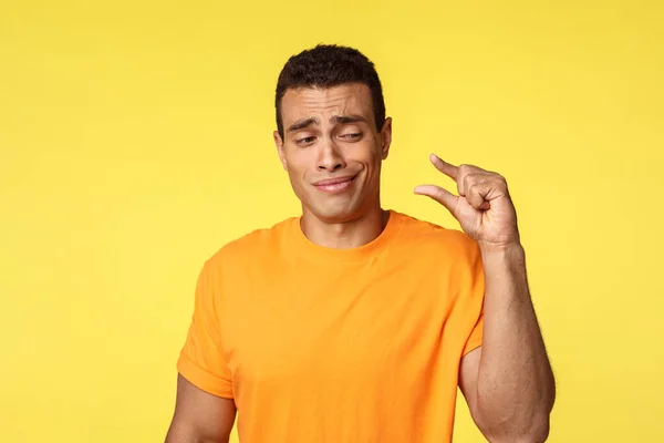 Cético e desapontado jovem bonito homem em camiseta laranja, mostrando algo pequeno, discutindo minúsculo objeto sorrir com desdém e olhar os dedos sem impressionar, de pé fundo amarelo — Fotografia de Stock