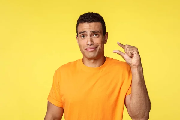 Disappointed and skeptical handsome masculine man in orange t-shirt cringe feeling awkward and indecisive, asking tiny offer, showing something small or little with gesture, yellow background — Stock Photo, Image