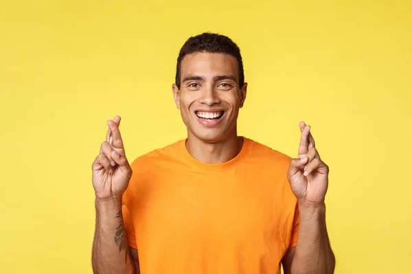 Close-up confiante, esperançoso, determinado jovem em camiseta laranja, assegurado que ele vai ganhar, dedos cruzados boa sorte, sorrindo feliz, auto-confiante receber elogios e feedback positivo, fundo amarelo — Fotografia de Stock