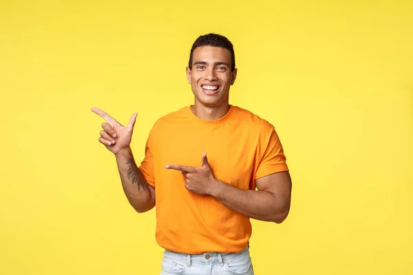 Cheerful modern man with tattooed arm, pointing left as promoting online store, invite guest join party, smiling joyfully, recommend try something, give advice what place visit, yellow background — Stock Photo, Image