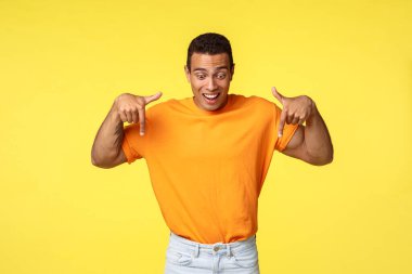 Heyecanlı, yakışıklı, İspanyol asıllı, erkeksi adam, turuncu tişört, beyaz pantolon, şaşkınlıktan kaşlarını kaldır, neşeyle gülümse ve şaşırmış bir şekilde aşağı bak.