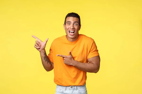 Cheerful excited man with tattooed arm in orange t-shirt, smiling astonished telling something amazing happened pointing left to promote exciting event, advice see yourself, yellow background — Stock Photo, Image