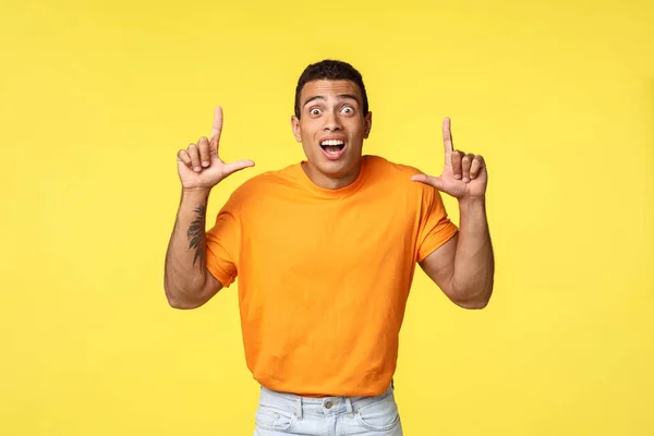 Fascinado, emocionado joven caucásico hombre en camiseta naranja, cámara de mirada asombrado e impresionado, apuntando hacia arriba como ver oferta increíble promo, de pie fondo amarillo sorprendido . — Foto de Stock