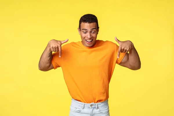 Beau garçon hispanique masculin excité, porter un t-shirt orange, pantalon blanc, lever les sourcils de l'étonnement et de l'impression, sourire joyeusement, pointant et regardant vers le bas surpris — Photo
