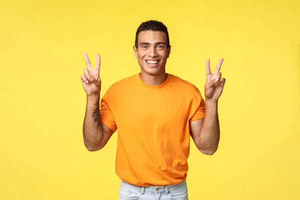 Guapo joven alegre en camiseta naranja, sonriendo optimista, hacer gesto de paz o citas signo, de pie fondo amarillo, expresar positividad y optimismo, de pie fondo amarillo — Foto de Stock