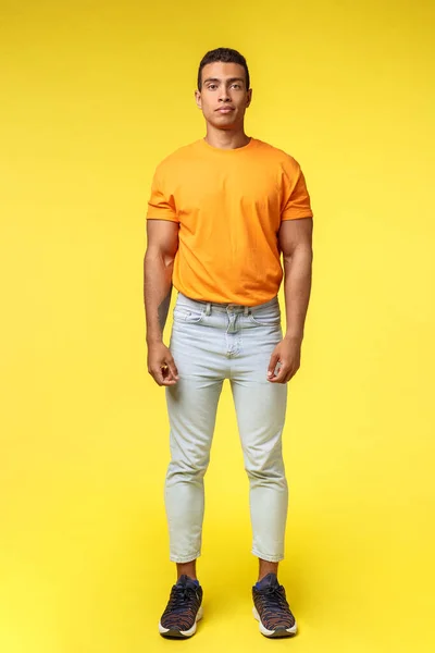 Full-length vertical shot masculine hipster guy, hispanic ethnicity, standing casually yellow background, wear stylish orange t-shirt, white pants, look camera with no expression, slightly smiling — Stock Photo, Image