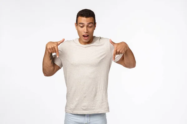 Fasciné et étonné, bel homme musclé hispanique sans voix en t-shirt blanc, retenant souffle de fascination et d'admiration, regard surpris vers le bas, pointant vers le bas promo, fond blanc — Photo