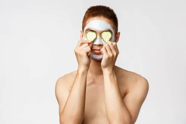 Szépség, bőr és spa koncepció. Portré vicces jóképű ázsiai férfi áll meztelenül a fehér háttér, visel uborka darab a szemek, alkalmazni arc maszk kezelésére akne és foltok — Stock Fotó