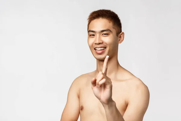 Schönheit, Menschen und Freizeitkonzept. Glücklich gut aussehender asiatischer Mann mit nacktem Oberkörper, zeigt Creme auf der Fingerkuppe und lächelt, trägt Hautpflegeprodukt auf das Gesicht auf, pflegt Flecken, weißer Hintergrund — Stockfoto