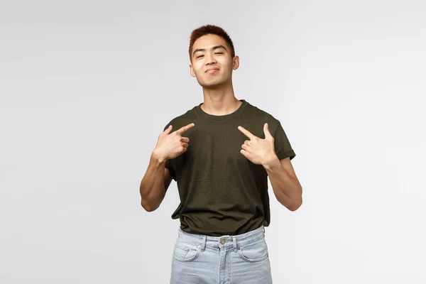 Portret dumnego i chełpliwego młodego azjatyckiego macho, pokazującego swoje wielkie ego, pokazującego się z uśmiechem na twarzy, aparatem fotograficznym, mówiącego o osobistych osiągnięciach, szarym tle — Zdjęcie stockowe