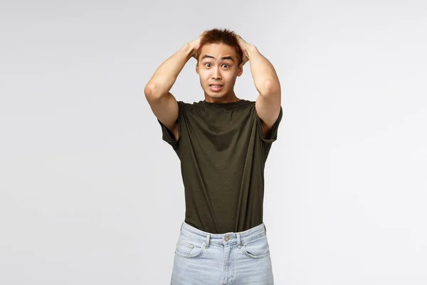 Portrait of alarmed young asian male student got in trouble, holding hands on head and staring camera indecisive, dont know what do, concerned standing grey background — Stock Photo, Image