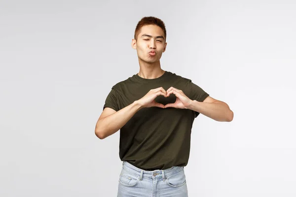 Mensen, emoties en levensstijl concept. Schattige en domme mannelijke student, aziatische man knipogen brutaal en flirterig, pruilen of vouwen lippen voor kus en tonen hart over borst, uiten sympathie en liefde — Stockfoto