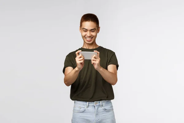 Technologie, online lifestyle en communicatie concept. Blij tevreden jonge aziatische man met behulp van mobiele telefoon, glimlachend vrolijk als het spelen van nieuwe smartphone app, grijns tevreden, grijze achtergrond — Stockfoto