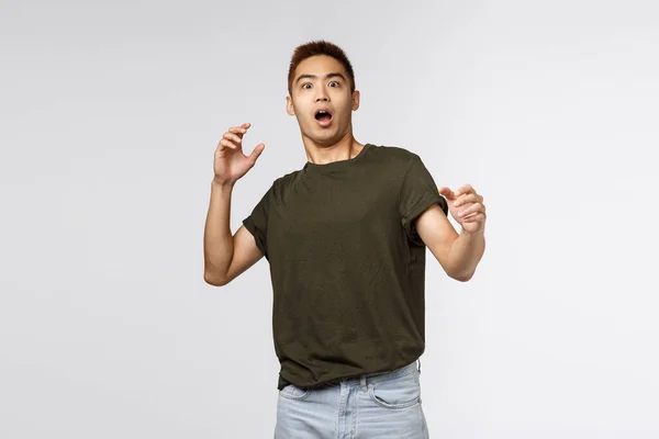 Retrato de alarmado y asustado, sorprendido chico asiático jadeando, paso atrás y levantando las manos asustado, sobresaltado mirando algo aterrador, de pie con asombro sobre fondo gris — Foto de Stock