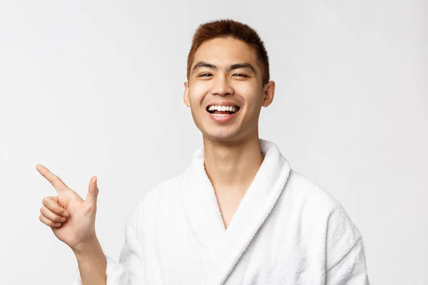 Beauty, Wellness- und Freizeitkonzept. Porträt eines fröhlichen asiatischen Mannes im Bademantel, genießt den Urlaub, zeigt mit dem Finger nach links, zeigt den Weg, lacht und lächelt zufrieden, steht weißen Hintergrund — Stockfoto
