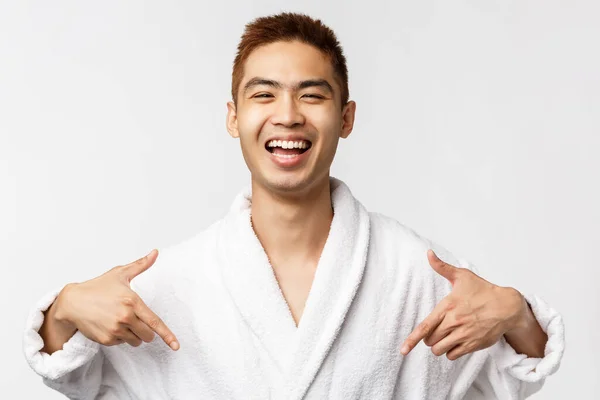 Krása, lázně a volný čas koncept. Portrét spokojený, šťastný asijský muž v županu ukazuje na sebe a široce se usmívá, mluví vlastní úspěch, osobní úspěch, bílé pozadí — Stock fotografie