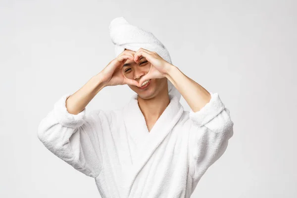 Conceito de beleza, spa e lazer. Adorável asiático homem em roupão de banho e toalha de banho, mostrar ouvir sinal sobre o rosto e sorrindo através dele, ficar otimista, desfrutar de tempo relaxante no hotel, fundo branco — Fotografia de Stock