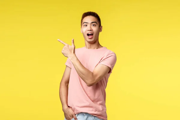 Lidé, různé emoce a životní styl. Vzrušený, šťastný asijský muž v růžovém tričku ukazující cestu, ukazující prstem v levém horním rohu a usmívající se ohromeně, doporučujeme navštívit obchod nebo kavárnu — Stock fotografie